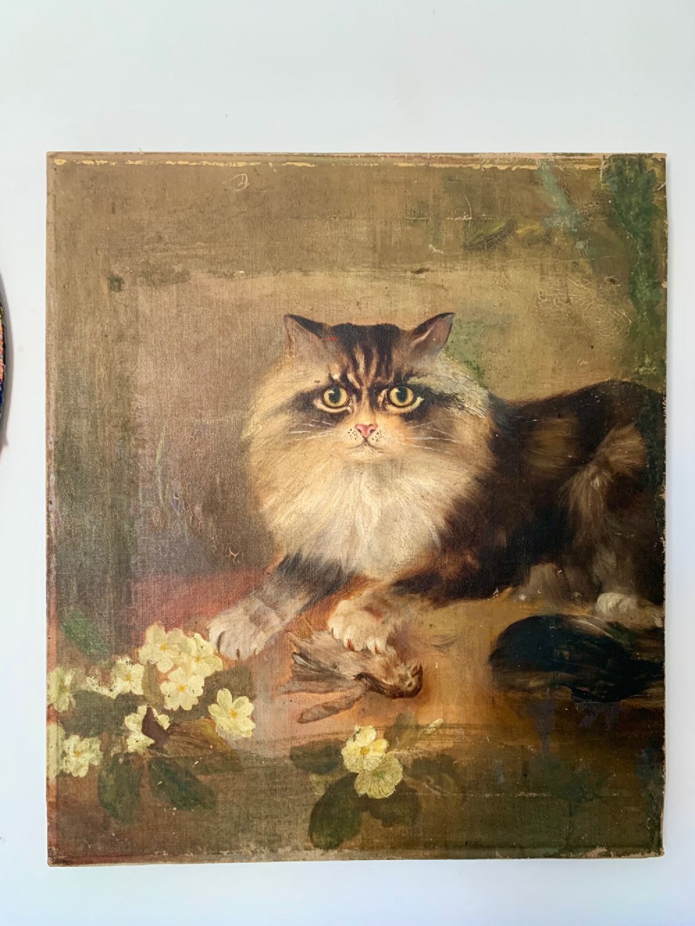 A 19th-century naive school oil painting depicting a fluffy longhaired cat holding a small bird, set beside pale cream flowers against a warm brown background. View II