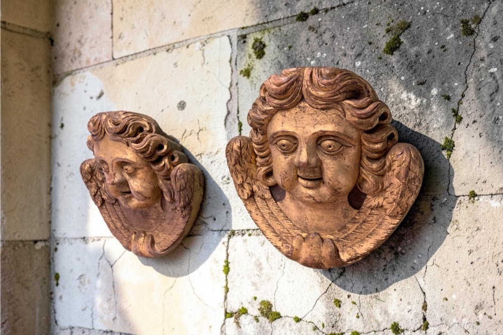A pair of French carved wooden church angel heads featuring curly hair and sculpted wings framing the face with remnants of gilded paint.