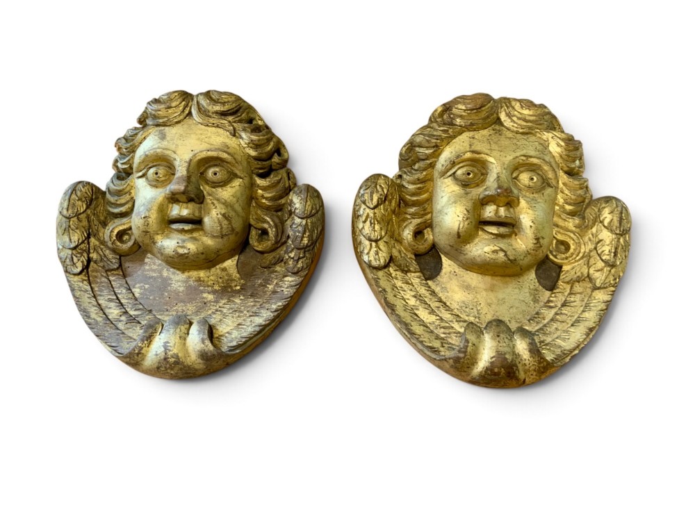 A pair of French carved wooden church angel heads featuring curly hair and sculpted wings framing the face with remnants of gilded paint. View II