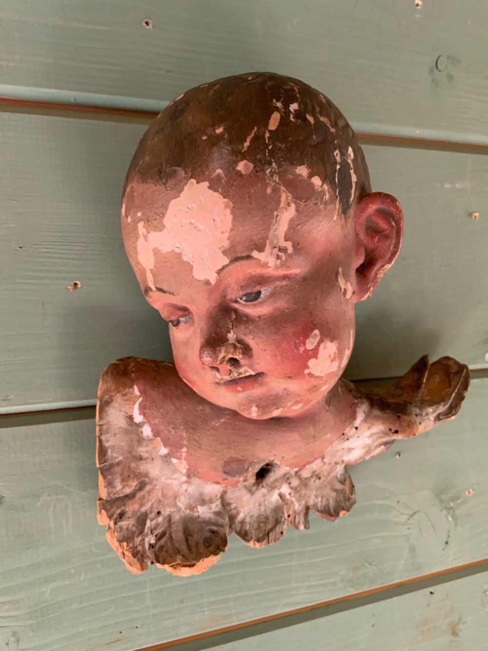 An early 18th-century carved wooden putto head featuring distressed polychrome paint and stylized winged detailing.