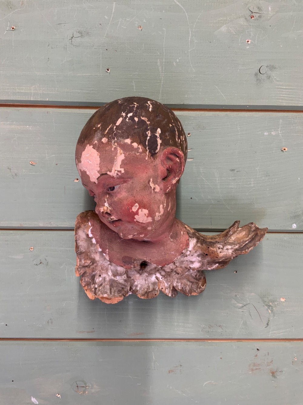 An early 18th-century carved wooden putto head featuring distressed polychrome paint and stylized winged detailing. View II