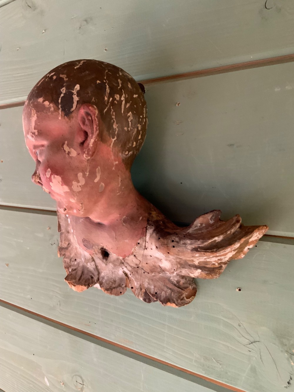 An early 18th-century carved wooden putto head featuring distressed polychrome paint and stylized winged detailing. View IV