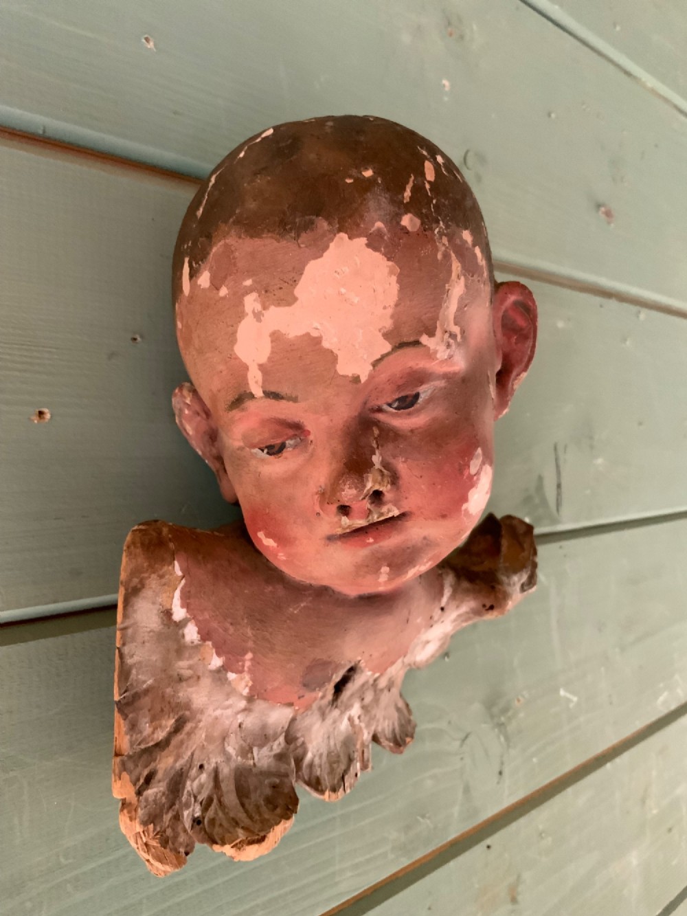 An early 18th-century carved wooden putto head featuring distressed polychrome paint and stylized winged detailing. View V