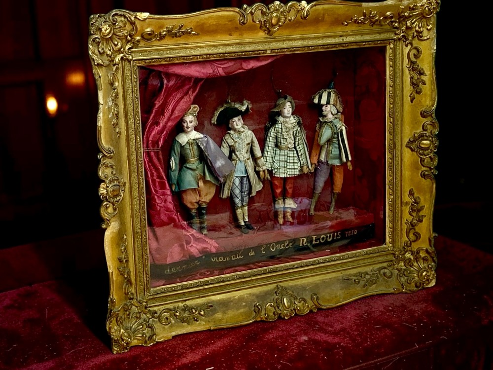 An antique gilt-framed shadow box diorama depicting four miniature costumed theatrical figures against a crimson draped fabric backdrop. View II
