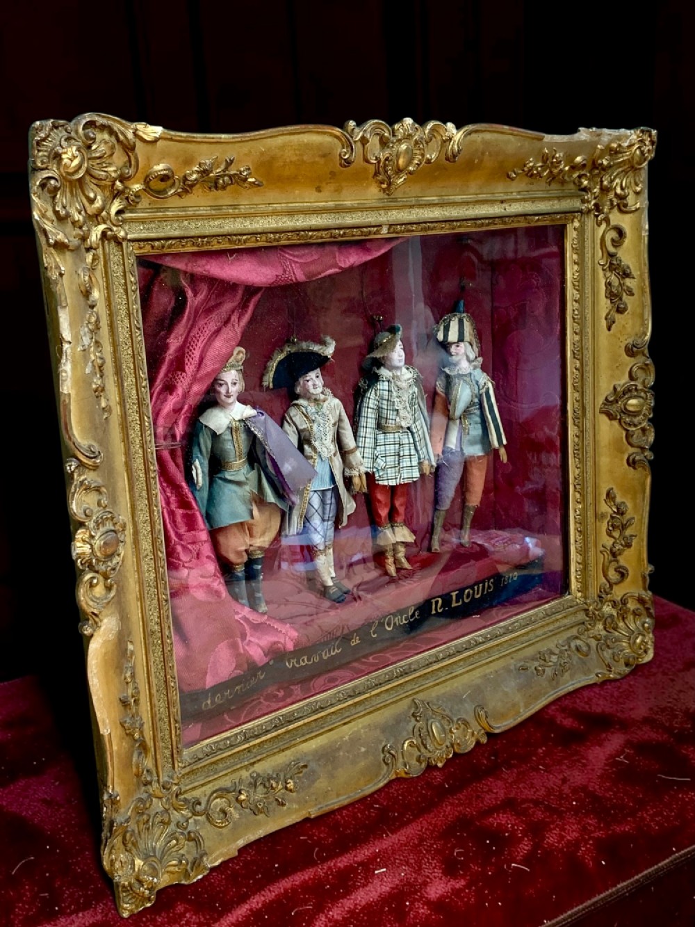 An antique gilt-framed shadow box diorama depicting four miniature costumed theatrical figures against a crimson draped fabric backdrop. View III