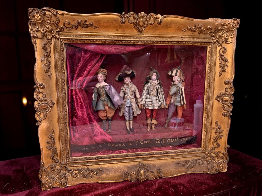 An antique gilt-framed shadow box diorama depicting four miniature costumed theatrical figures against a crimson draped fabric backdrop. View IV