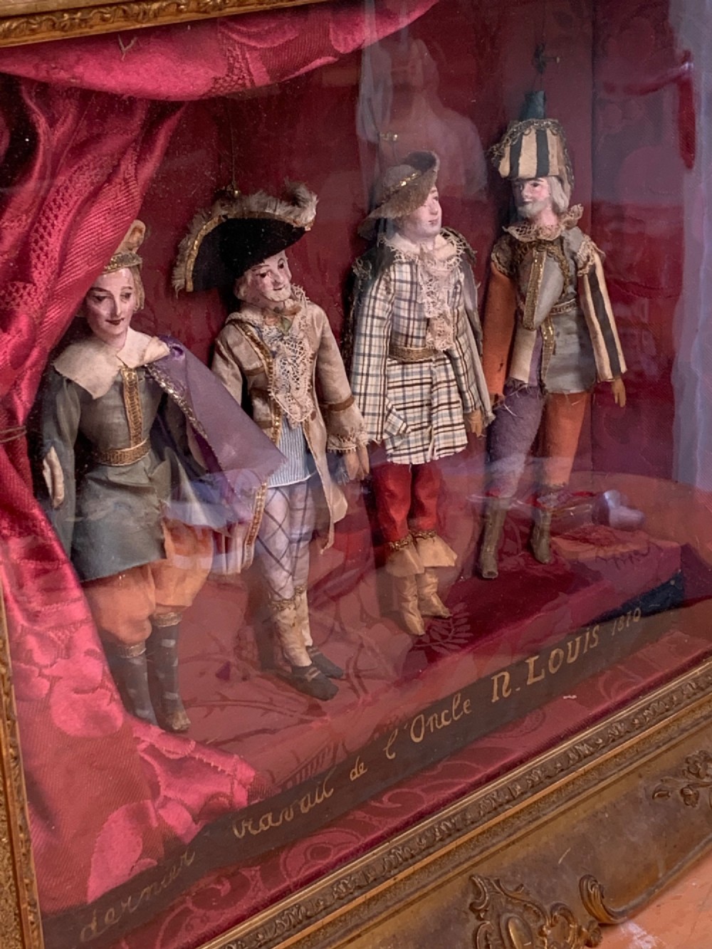 An antique gilt-framed shadow box diorama depicting four miniature costumed theatrical figures against a crimson draped fabric backdrop. View V