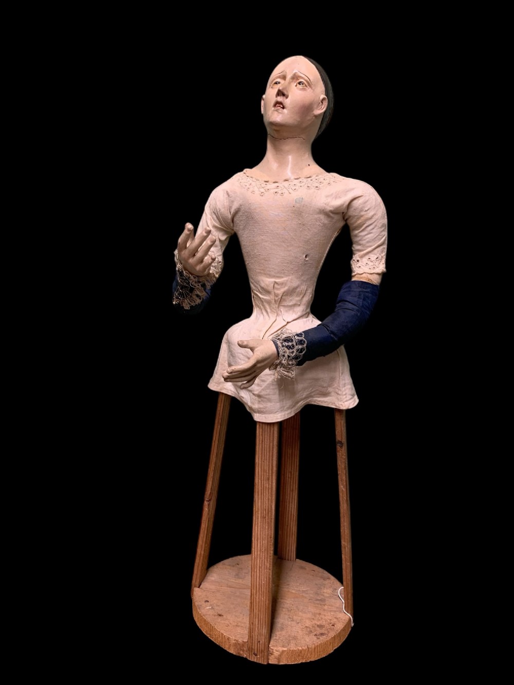 A 19th-century processional mannequin with carved polychrome head and hands, dressed in cream fabric with dark blue sleeves, standing on a four-legged wooden cage base.