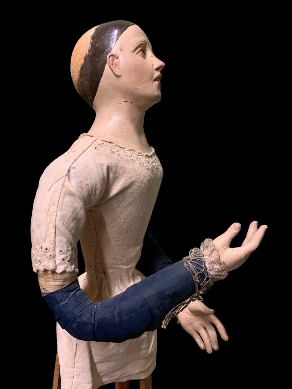 A 19th-century processional mannequin with carved polychrome head and hands, dressed in cream fabric with dark blue sleeves, standing on a four-legged wooden cage base. View IV