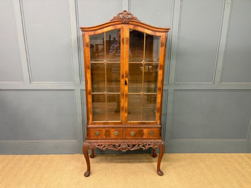 fig 1. An antique Queen Anne style glazed display cabinet in warm brown wood featuring arched doors with glass panes, drawers with brass handles, and carved cabriole legs.