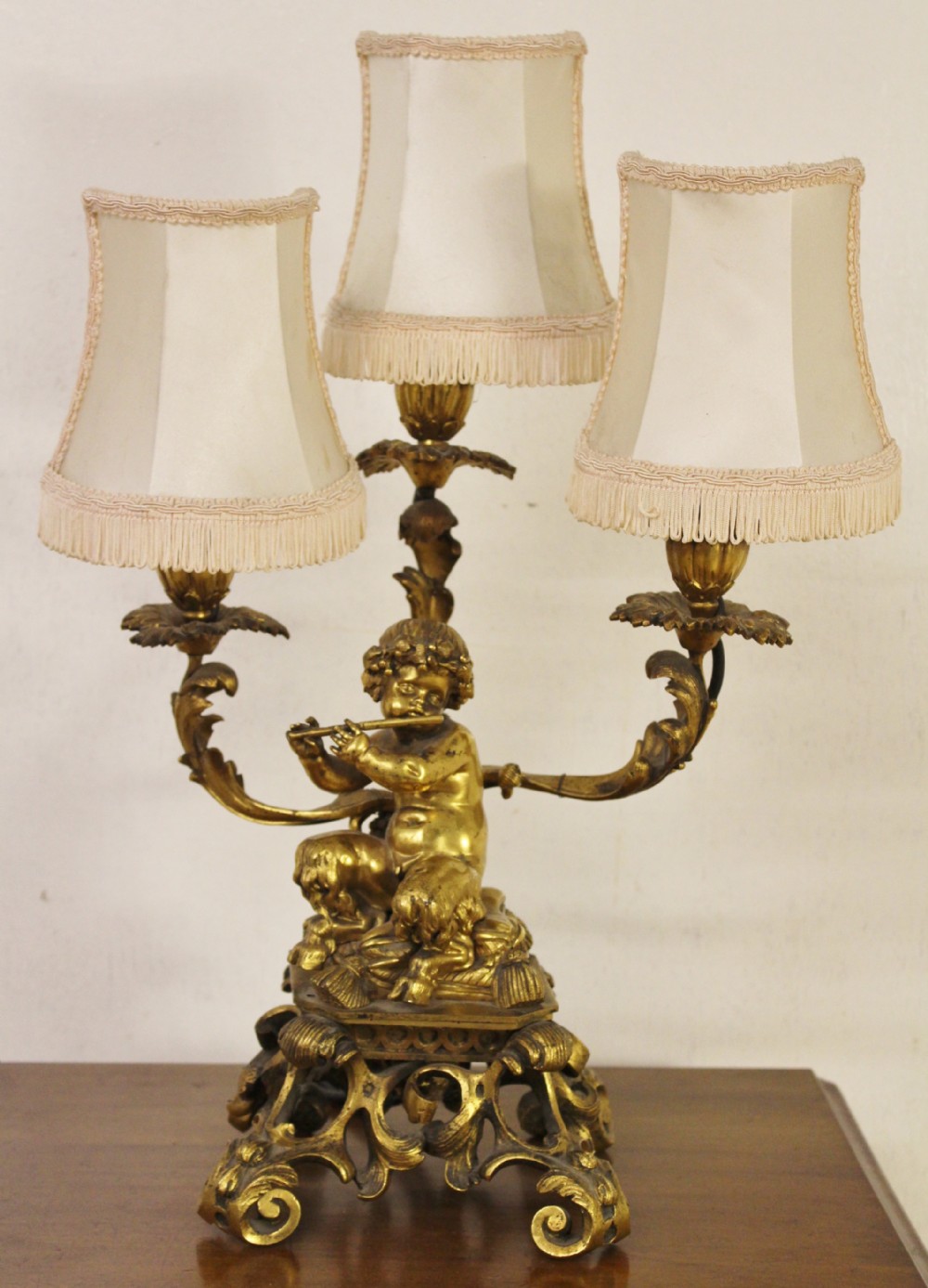 A gilt bronze table lamp featuring a central sculptural cherub figure playing a flute, supporting three scrolling light arms with cream fringed shades.
