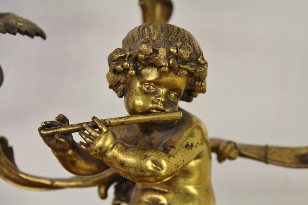 A gilt bronze table lamp featuring a central sculptural cherub figure playing a flute, supporting three scrolling light arms with cream fringed shades. View III