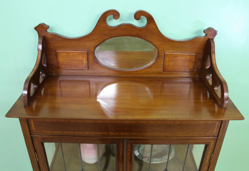 An Arts and Crafts inlaid mahogany display cabinet featuring leaded glass doors, a shaped gallery top with an oval mirror, and slender tapered legs. View II
