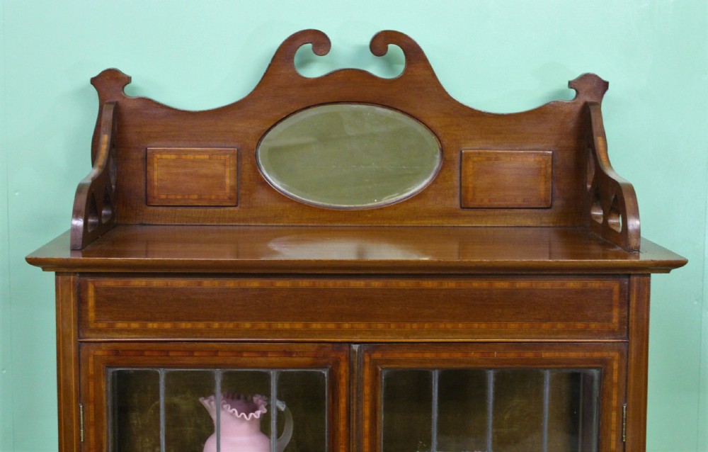 An Arts and Crafts inlaid mahogany display cabinet featuring leaded glass doors, a shaped gallery top with an oval mirror, and slender tapered legs. View VII