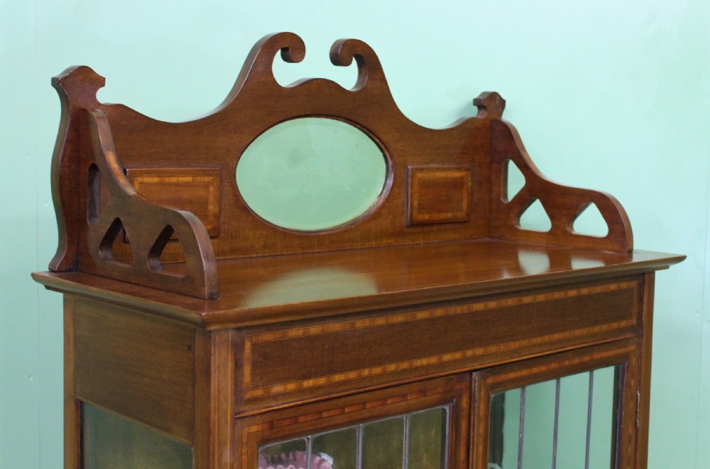 An Arts and Crafts inlaid mahogany display cabinet featuring leaded glass doors, a shaped gallery top with an oval mirror, and slender tapered legs. View XIII