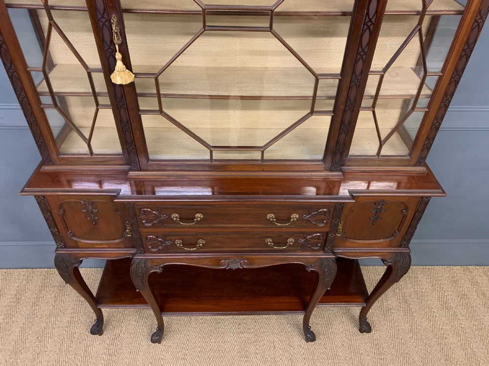 A carved dark mahogany display cabinet featuring three astragal glazed doors above drawers and cabriole legs on an undertier shelf. View VI