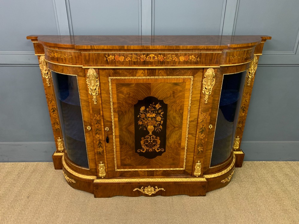 A Victorian burr walnut credenza featuring floral marquetry inlays, gilt bronze ormolu mounts, a central panelled door, and curved glazed side cabinets.