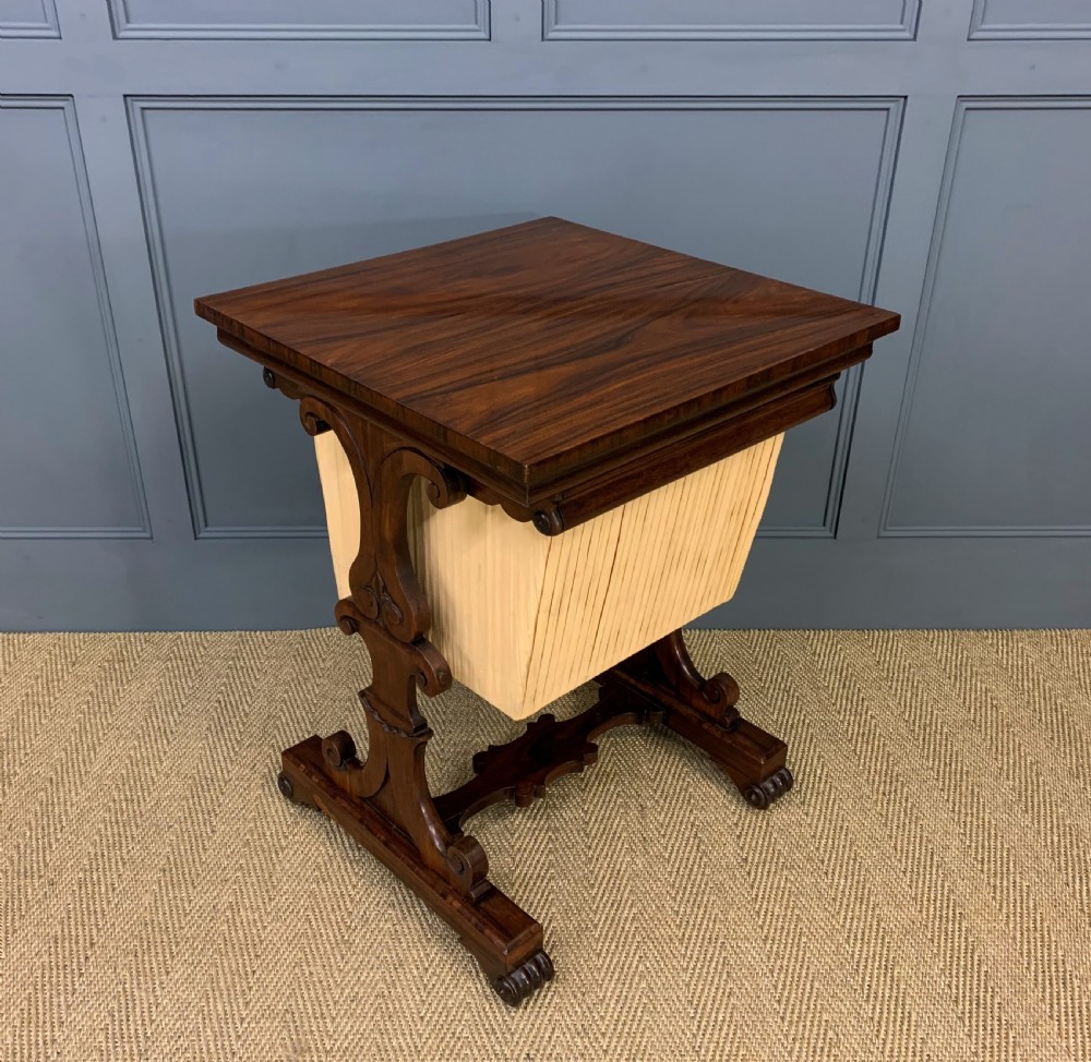 A William IV rosewood work table featuring a figured square top, pleated cream fabric basket, and carved scroll supports.