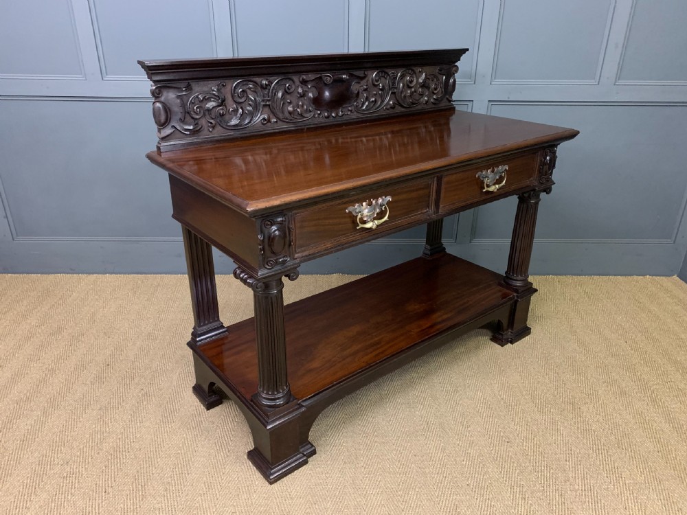 A Victorian mahogany buffet featuring a carved scroll backboard, two frieze drawers with brass handles, fluted support columns, and an open lower tier.