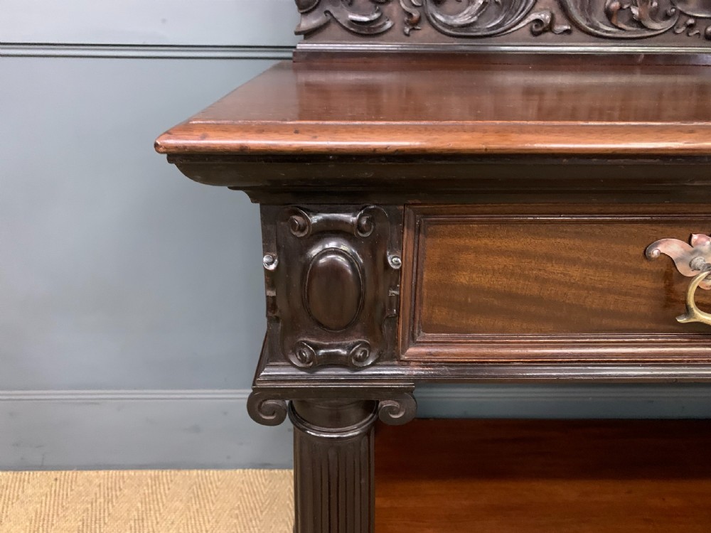 A Victorian mahogany buffet featuring a carved scroll backboard, two frieze drawers with brass handles, fluted support columns, and an open lower tier. View VII