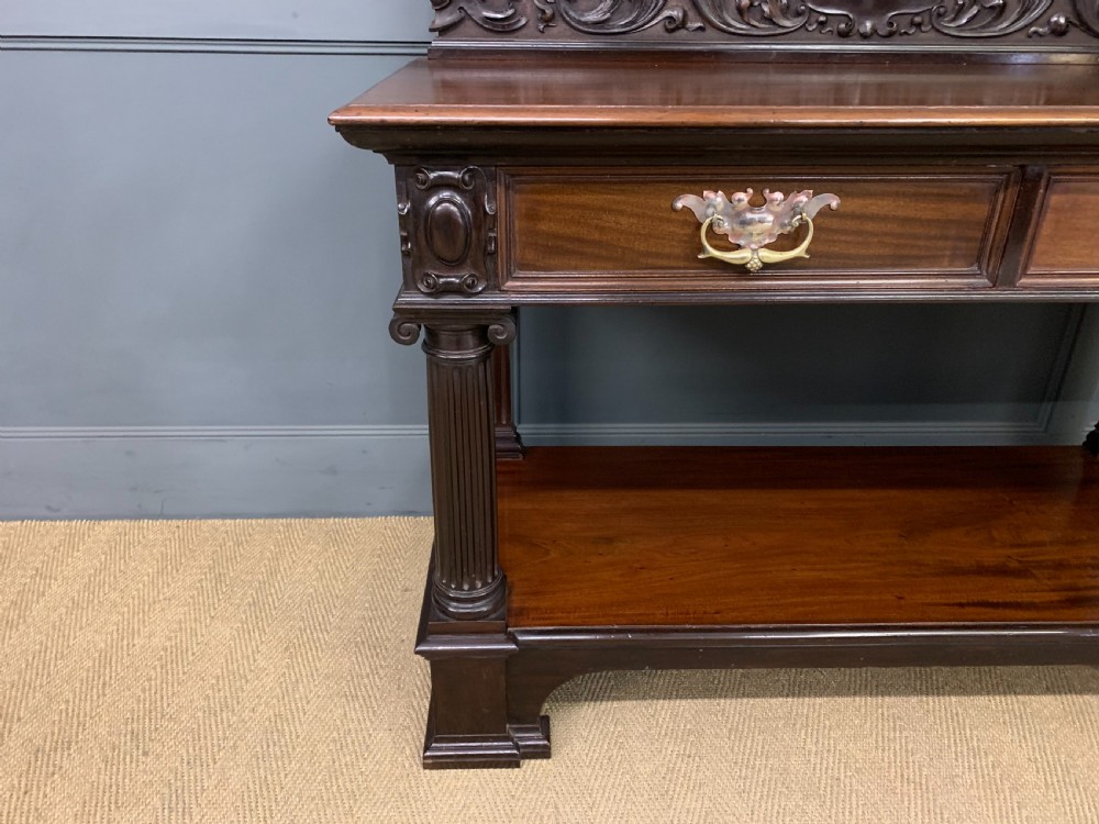A Victorian mahogany buffet featuring a carved scroll backboard, two frieze drawers with brass handles, fluted support columns, and an open lower tier. View X