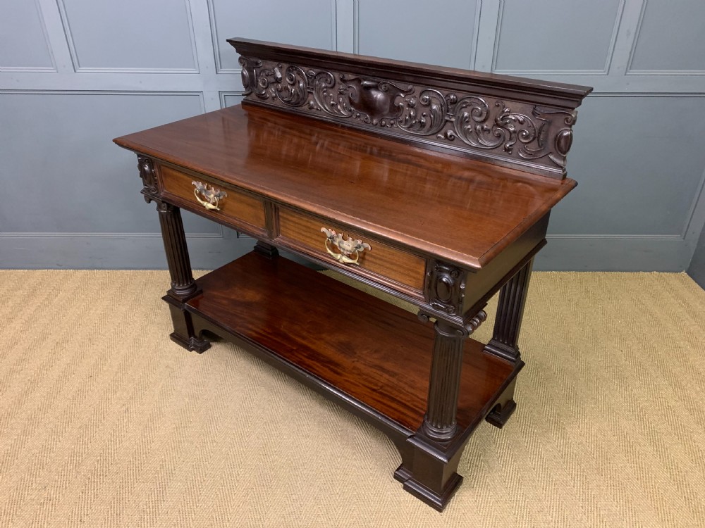 A Victorian mahogany buffet featuring a carved scroll backboard, two frieze drawers with brass handles, fluted support columns, and an open lower tier. View XV