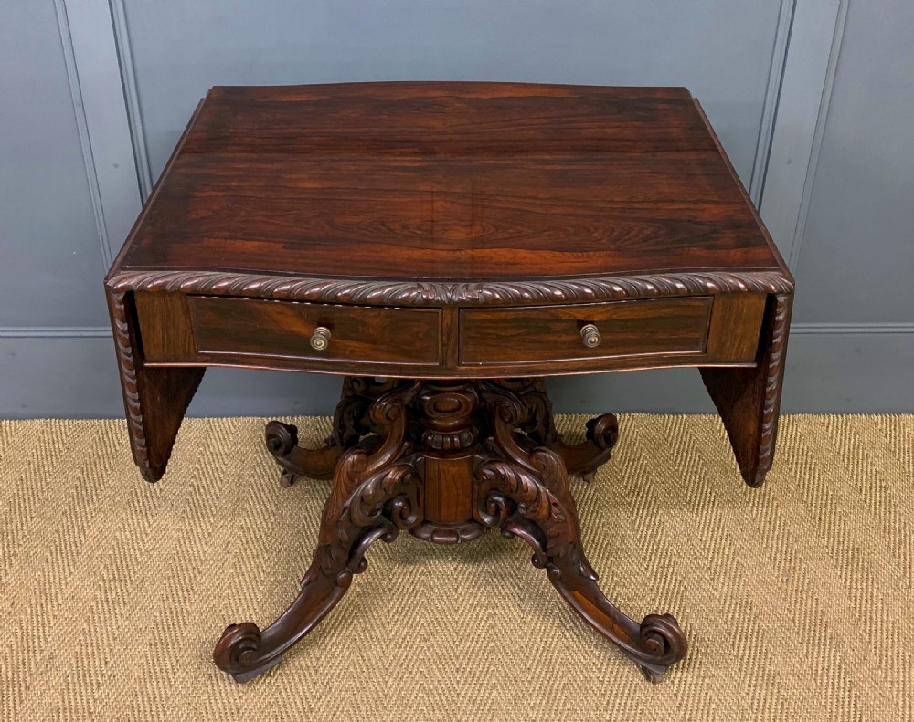 A 19th-century dark rosewood sofa table featuring drop leaves, two front frieze drawers, and an intricately carved central pedestal on scroll legs.