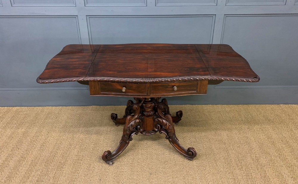 A 19th-century dark rosewood sofa table featuring drop leaves, two front frieze drawers, and an intricately carved central pedestal on scroll legs. View XIV