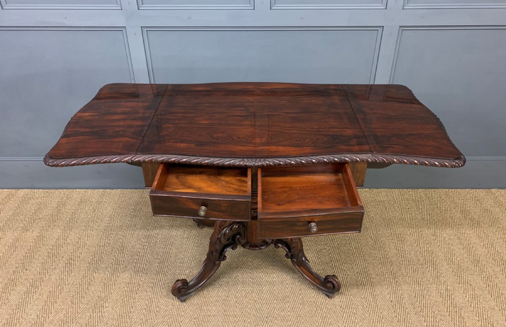 A 19th-century dark rosewood sofa table featuring drop leaves, two front frieze drawers, and an intricately carved central pedestal on scroll legs. View XIX