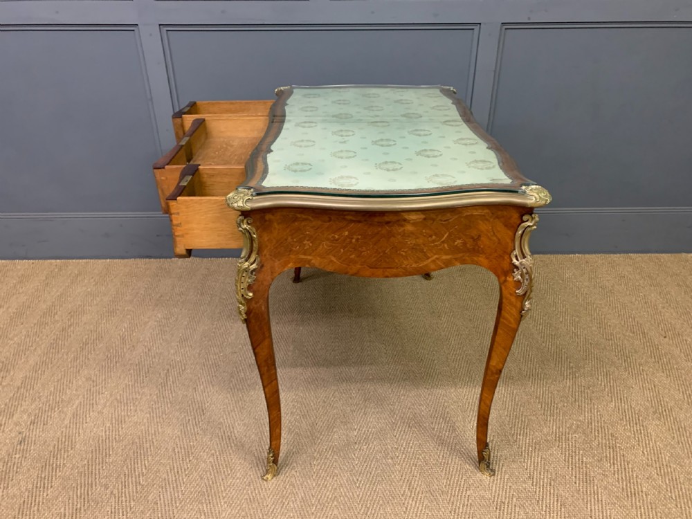 A French Louis XV style bureau plat featuring marquetry floral inlay, gilt-bronze mounts, cabriole legs, and a mint green patterned textile top under protective glass. View XV