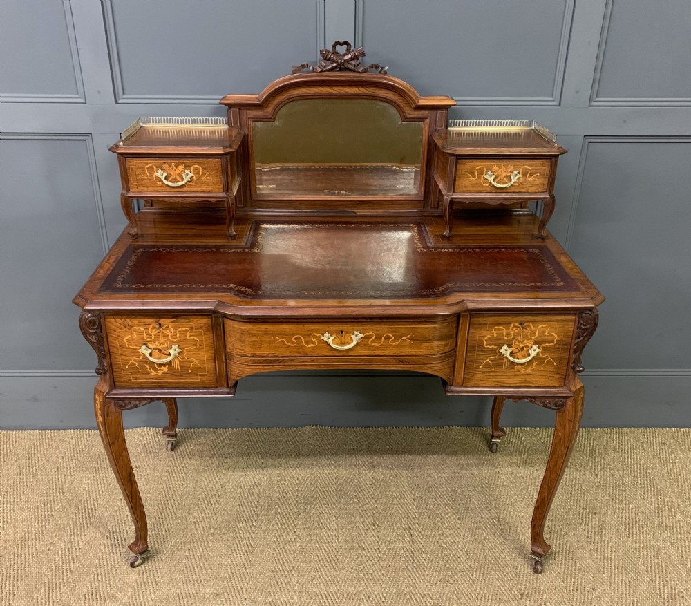A Victorian mahogany writing desk featuring floral marquetry inlay, a gold-tooled leather insert, a raised superstructure with two drawers and a central mirror, resting on cabriole legs with brass castors.