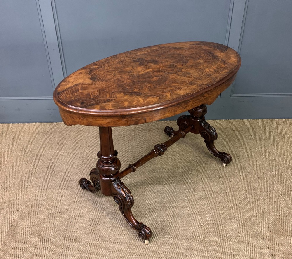 An oval Victorian burr walnut stretcher table featuring floral inlay, turned end columns, carved splayed legs, and ceramic castors.