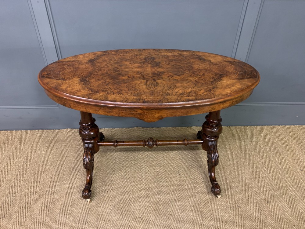 An oval Victorian burr walnut stretcher table featuring floral inlay, turned end columns, carved splayed legs, and ceramic castors. View II