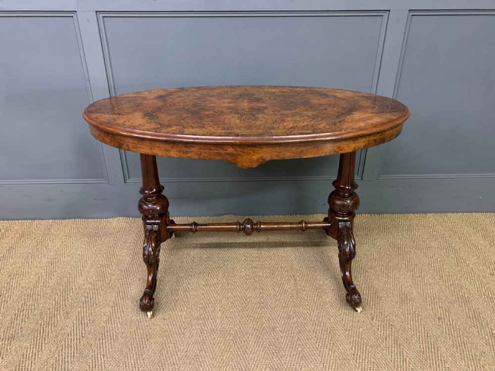 An oval Victorian burr walnut stretcher table featuring floral inlay, turned end columns, carved splayed legs, and ceramic castors. View III