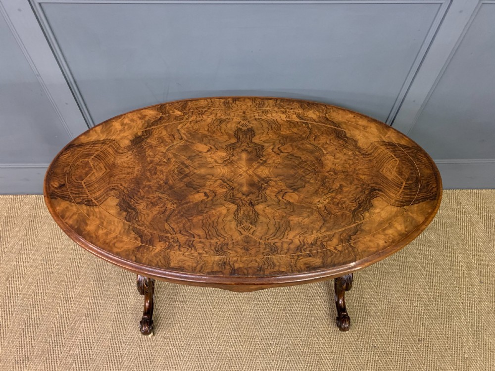 An oval Victorian burr walnut stretcher table featuring floral inlay, turned end columns, carved splayed legs, and ceramic castors. View IV