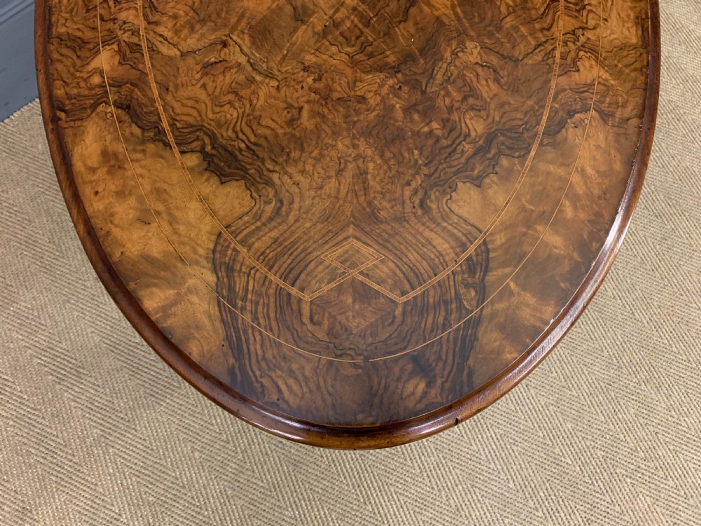An oval Victorian burr walnut stretcher table featuring floral inlay, turned end columns, carved splayed legs, and ceramic castors. View V