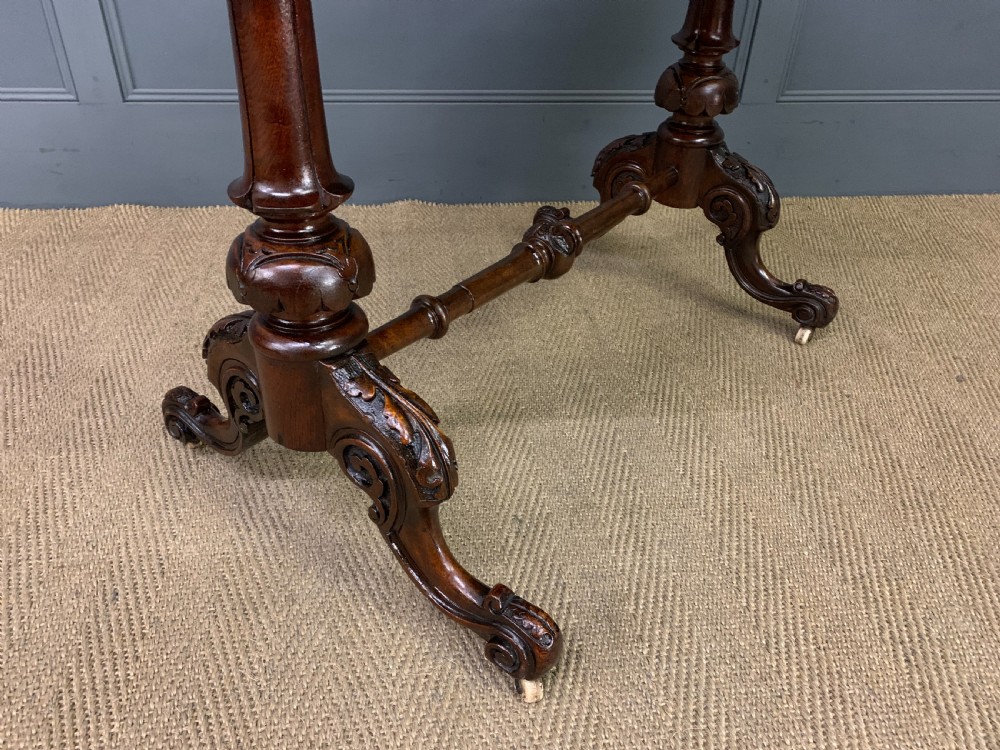 An oval Victorian burr walnut stretcher table featuring floral inlay, turned end columns, carved splayed legs, and ceramic castors. View VII