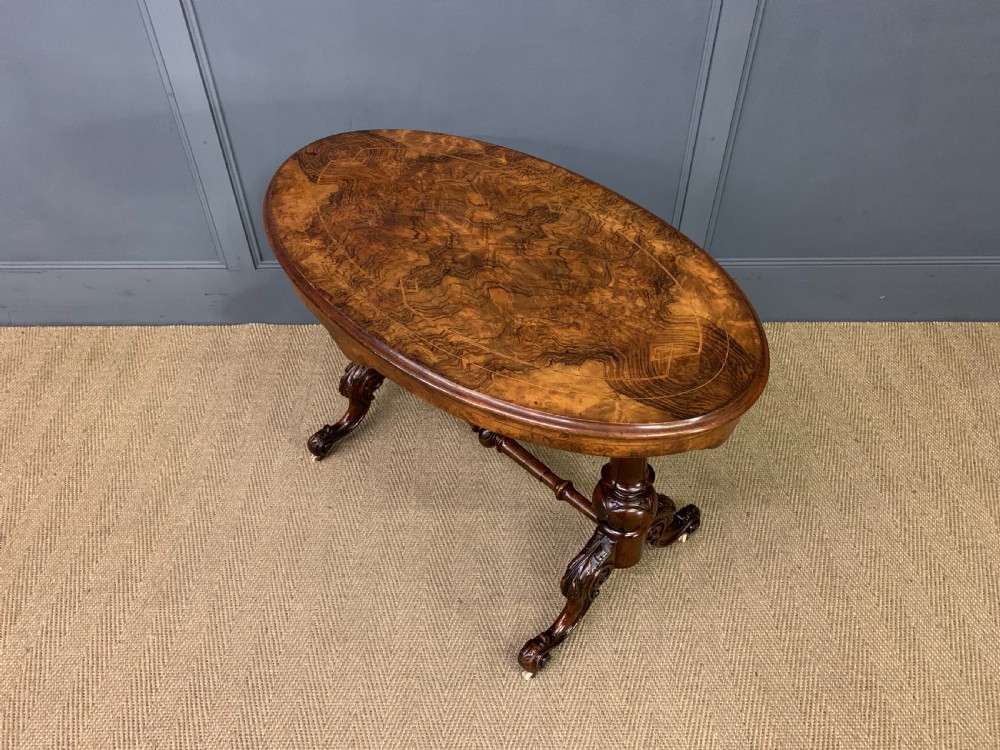 An oval Victorian burr walnut stretcher table featuring floral inlay, turned end columns, carved splayed legs, and ceramic castors. View IX