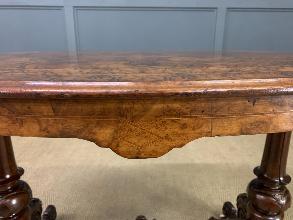 An oval Victorian burr walnut stretcher table featuring floral inlay, turned end columns, carved splayed legs, and ceramic castors. View X