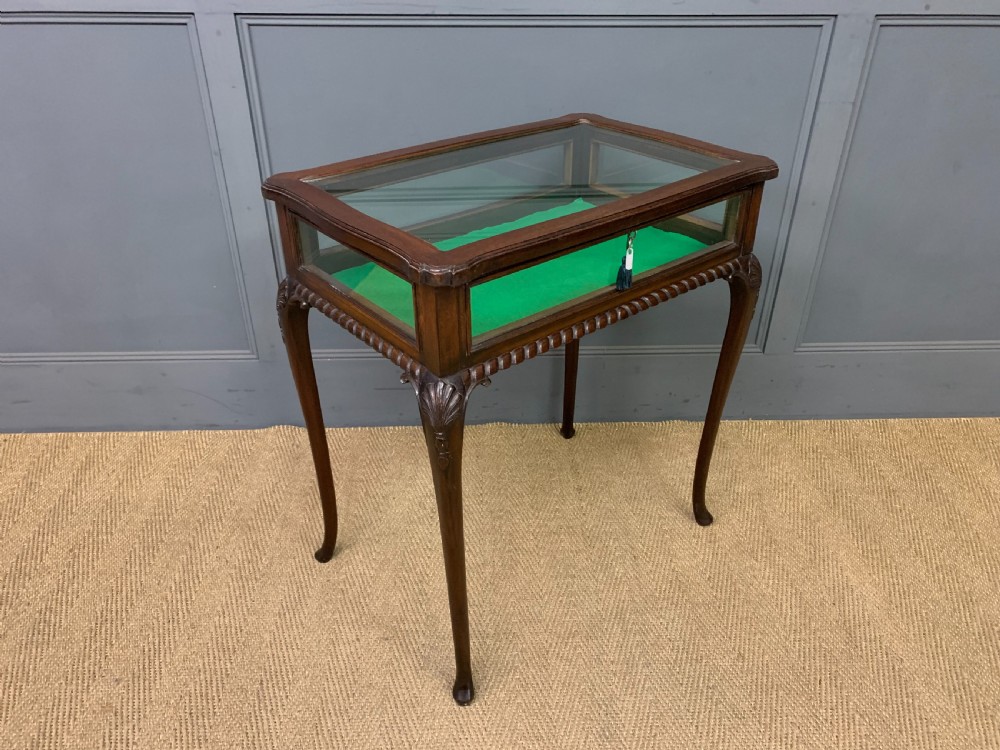 A mahogany bijouterie table featuring a glazed glass top and sides, green fabric interior lining, cabriole legs, and carved shell details on the knees.