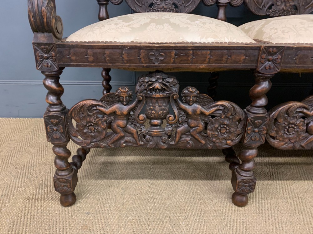 A 19th-century Carolean style walnut three-seat settee featuring barley twist columns, ornate openwork cherub carvings, and cream damask upholstered back panels and seats. View VI