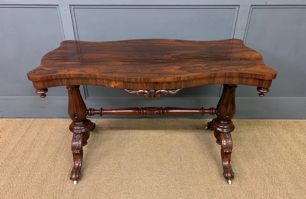 Victorian rosewood stretcher table featuring a serpentine top with figured grain, turned end supports, a carved central stretcher, and ceramic castors.