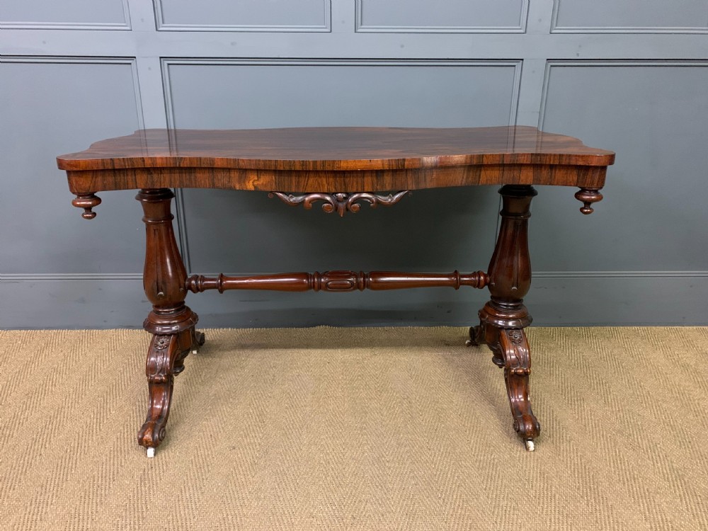 Victorian rosewood stretcher table featuring a serpentine top with figured grain, turned end supports, a carved central stretcher, and ceramic castors. View II