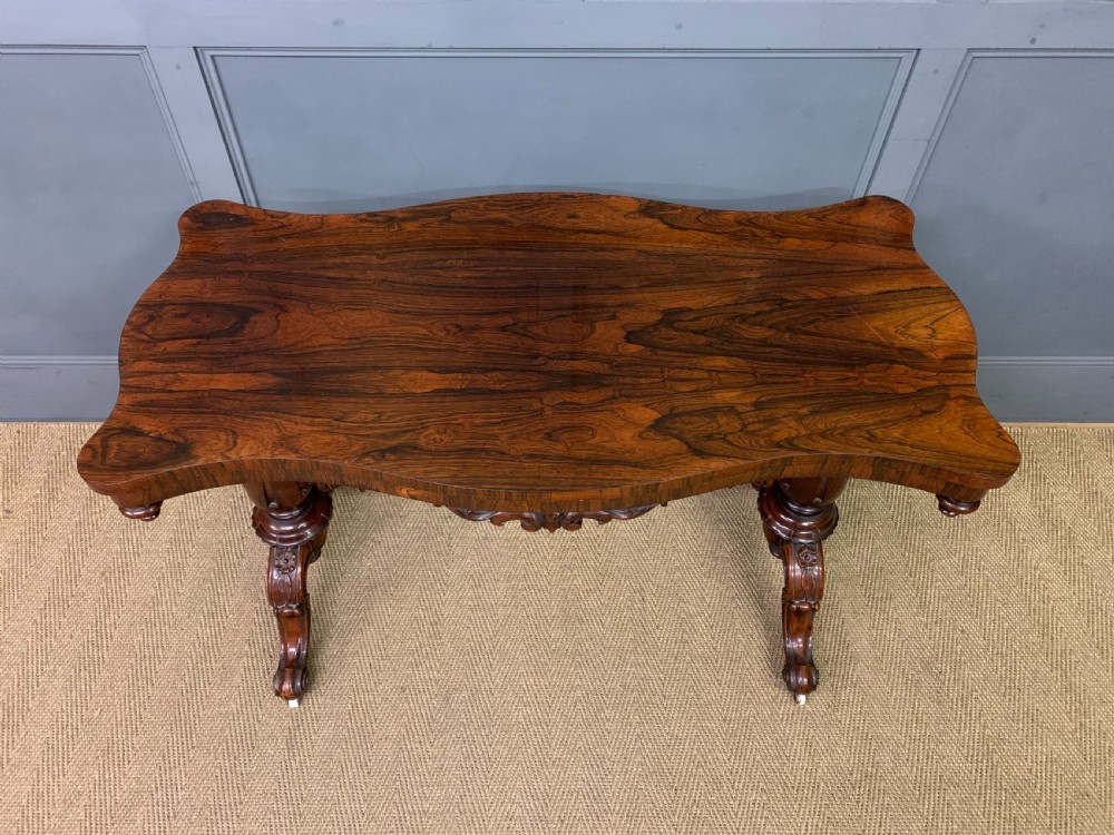 Victorian rosewood stretcher table featuring a serpentine top with figured grain, turned end supports, a carved central stretcher, and ceramic castors. View III