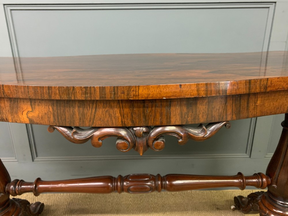 Victorian rosewood stretcher table featuring a serpentine top with figured grain, turned end supports, a carved central stretcher, and ceramic castors. View IV