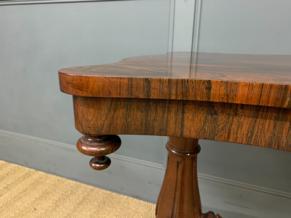 Victorian rosewood stretcher table featuring a serpentine top with figured grain, turned end supports, a carved central stretcher, and ceramic castors. View V