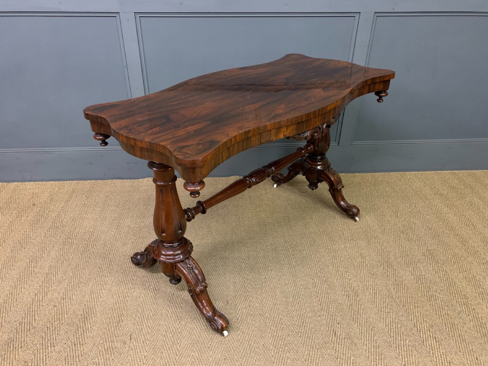 Victorian rosewood stretcher table featuring a serpentine top with figured grain, turned end supports, a carved central stretcher, and ceramic castors. View VI