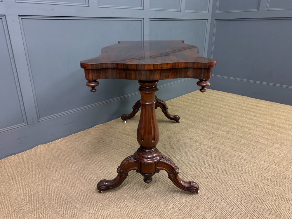 Victorian rosewood stretcher table featuring a serpentine top with figured grain, turned end supports, a carved central stretcher, and ceramic castors. View VII