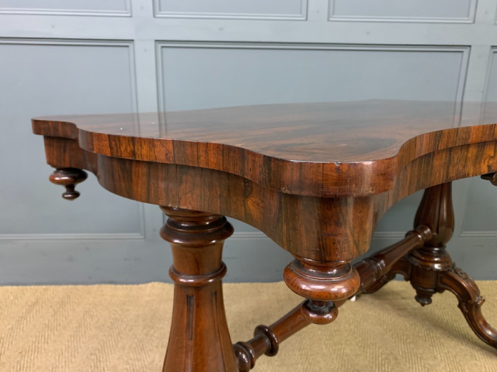 Victorian rosewood stretcher table featuring a serpentine top with figured grain, turned end supports, a carved central stretcher, and ceramic castors. View X