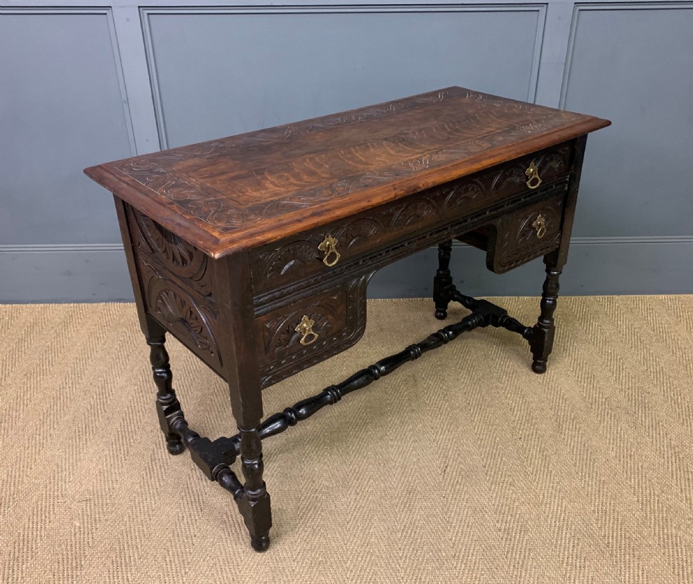 A dark carved oak side table featuring intricate lunette carvings, brass drop handles, three drawers, turned legs, and a stretcher base.
